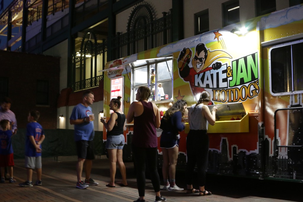 Guests at the Kate & Jan Hotdogs truck on a brick plaza at night outside Wrigley Field